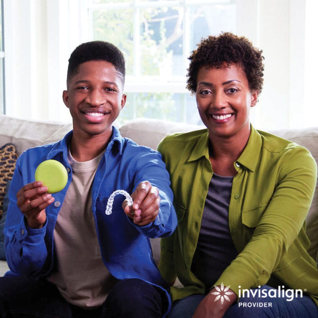 Two people smiling on a couch, one holding an Invisalign case and aligner, embodying the future of image orthodontics. The Invisalign Provider logo is clearly visible, highlighting the innovative shift from traditional braces to modern solutions delivered by expert orthodontists.