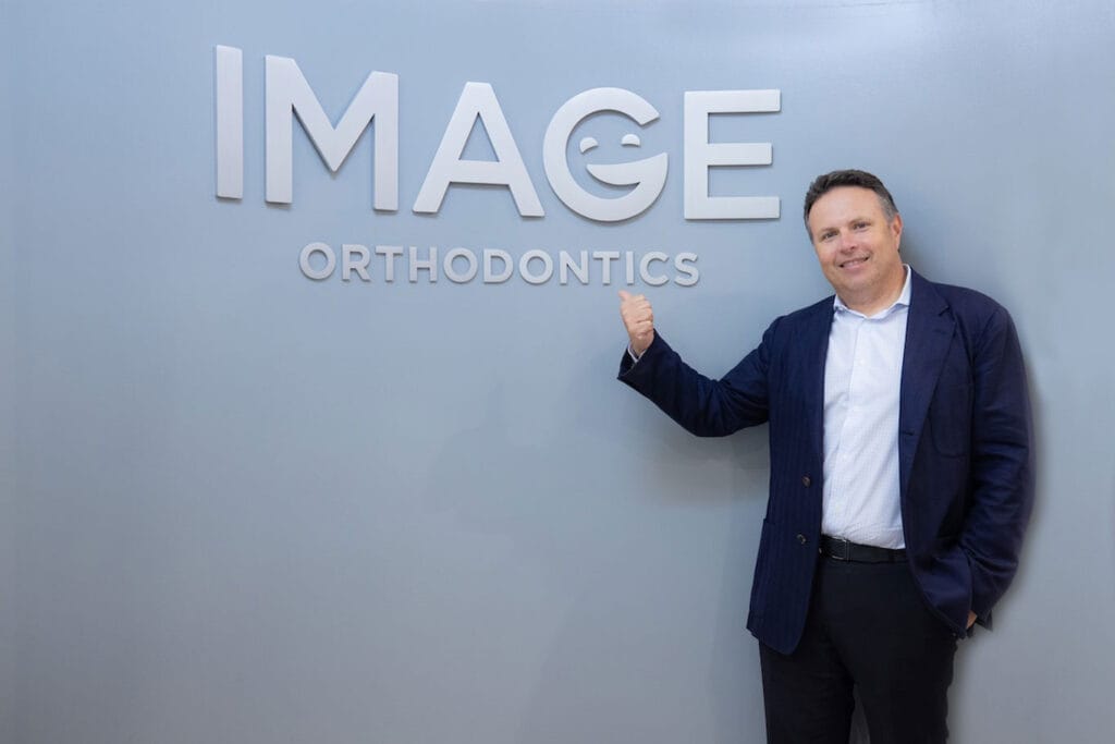 Dr. Yan Kalika in a suit stands smiling, pointing at a wall sign that reads "IMAGE ORTHODONTICS."