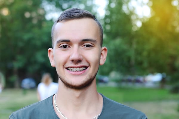 A young man with clear braces smiles in an outdoor setting, reflecting the expert touch of his orthodontist against a backdrop of blurred greenery.
