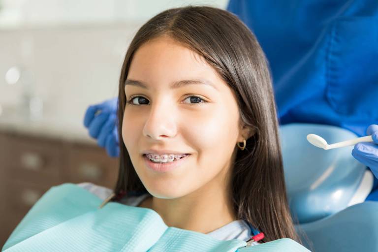 A person with braces or possibly Invisalign sits in a dental chair, wearing a dental bib. A dentist, visible partially, holds a dental tool nearby.