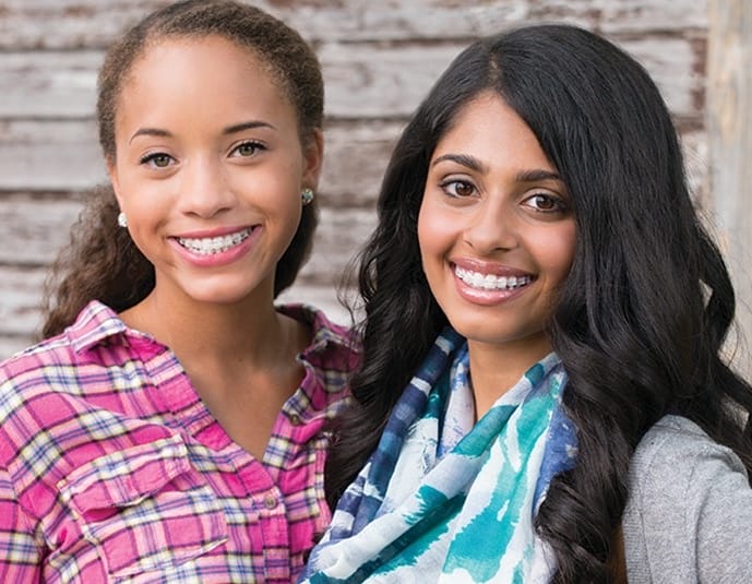 Two young ladies with clear braces flash bright smiles at the camera, standing proudly in front of a wooden background, showcasing the work of their orthodontist.