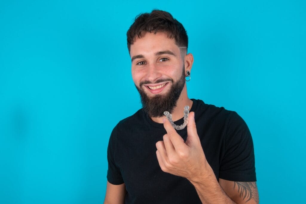 A bearded person holds clear aligners, smiling confidently against a blue background, showcasing the transformative magic of modern orthodontics.