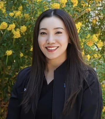 Dr. Vicky Chiu. A woman with long dark hair smiles in front of yellow flowers, wearing a black jacket, capturing the essence of joy that mirrors a visit to an orthodontist in Pleasant Hill.