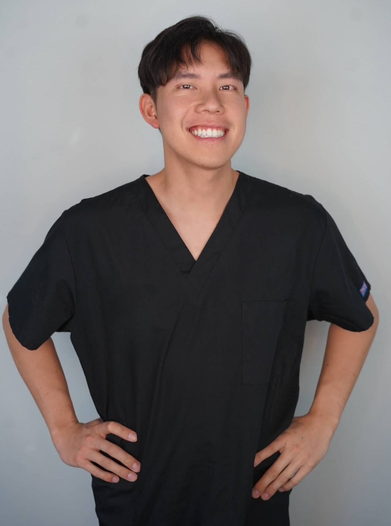 Dr. Ryan Gaw, DDS. A person smiling confidently, standing with hands on hips in a black V-neck shirt against a plain background, exuding the assurance of an orthodontist from Turlock.