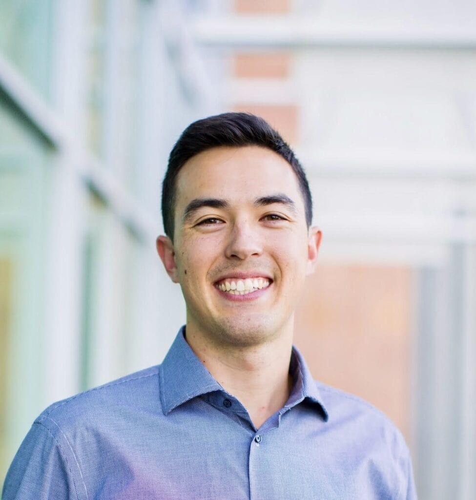 A man wearing a light blue button-up shirt smiles while standing in front of a modern glass building, reflecting the professionalism you'd expect from a Turlock orthodontist.