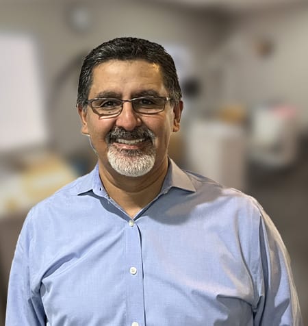 A man with glasses and a beard smiles, wearing a light blue shirt. The background is a blurred office setting, hinting at his role as an orthodontist in San Jose Berryessa.