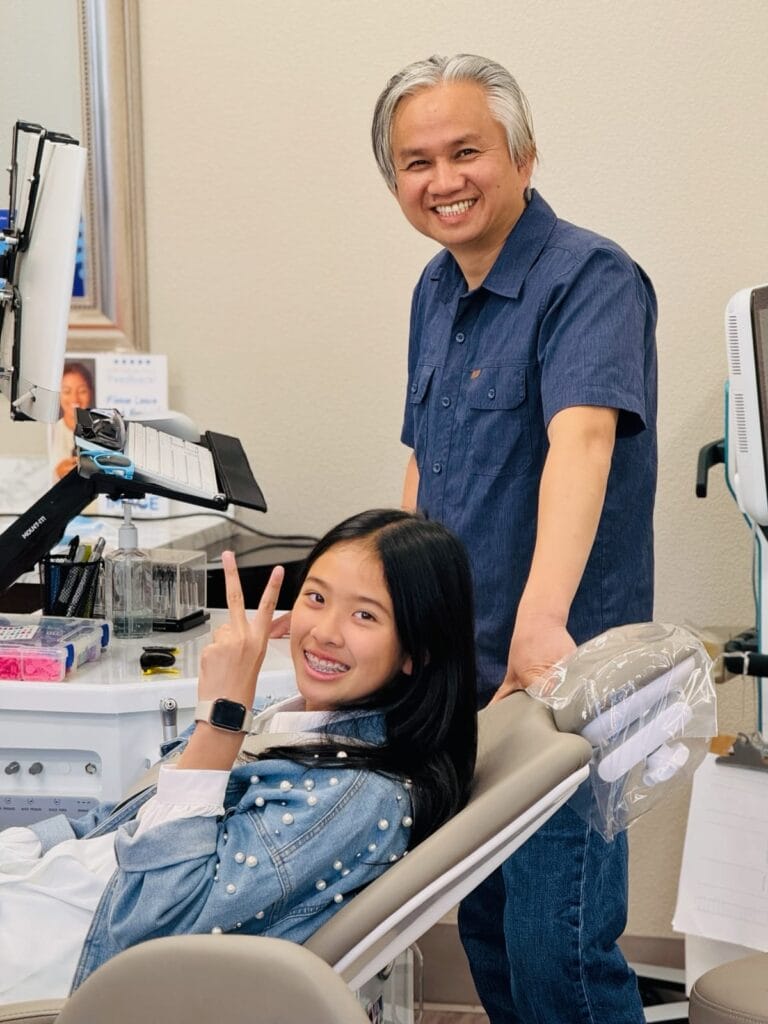 A girl with braces sits in a dental chair making a peace sign, while a Rocklin orthodontist stands beside her, smiling at the camera in the dental office.