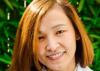 Dr. Gracia Cua, DMD, MScD. A person with a bright smile and short brown hair, wearing a light-colored top, stands against a leafy green backdrop. This lively scene captures the vibrant spirit often found in South San Francisco's community, reminiscent of the friendly atmosphere at your local orthodontist.