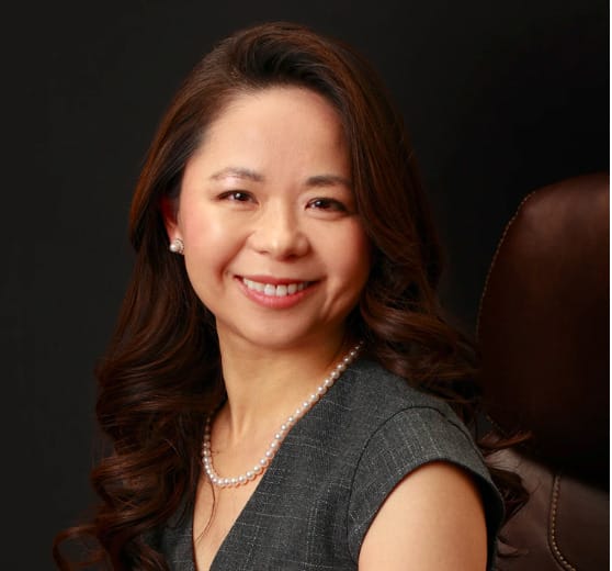 Dr. Melissa Bailey, DDS, MS. A smiling woman with long brown hair, wearing a gray outfit and pearl necklace, is seated against a dark background, reminiscent of the calming assurance of an experienced Pleasanton orthodontist.