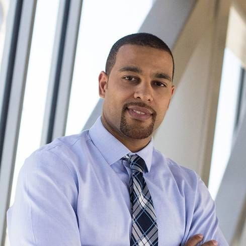 Dr. Michael Simontacchi-Gbologah. An orthodontist in a light blue shirt and plaid tie stands with arms crossed in front of large windows, embodying the commitment and care you can expect from a Fairfield orthodontist.