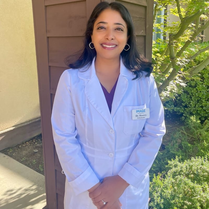 Dr. Shruti Shastry. A person in a white coat stands outdoors, smiling amidst the greenery and wooden structure. Nearby, the renowned San Jose Blossom Hill orthodontist offers expert care in a tranquil setting.