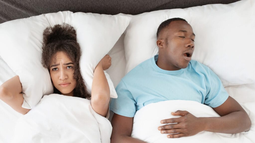 A woman covers her ears with a pillow looking annoyed while a man beside her sleeps with his mouth open, possibly snoring.