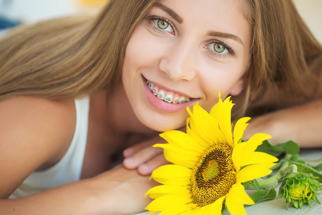 A cheerful individual with braces beams while lying on the ground beside a vibrant yellow sunflower, showcasing their radiant smile and confidence.