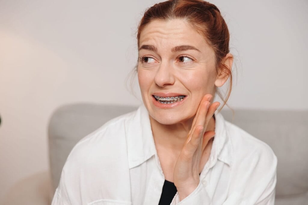 Woman with braces holding her jaw and appearing to be in pain, indicating discomfort possibly related to orthodontic treatment.