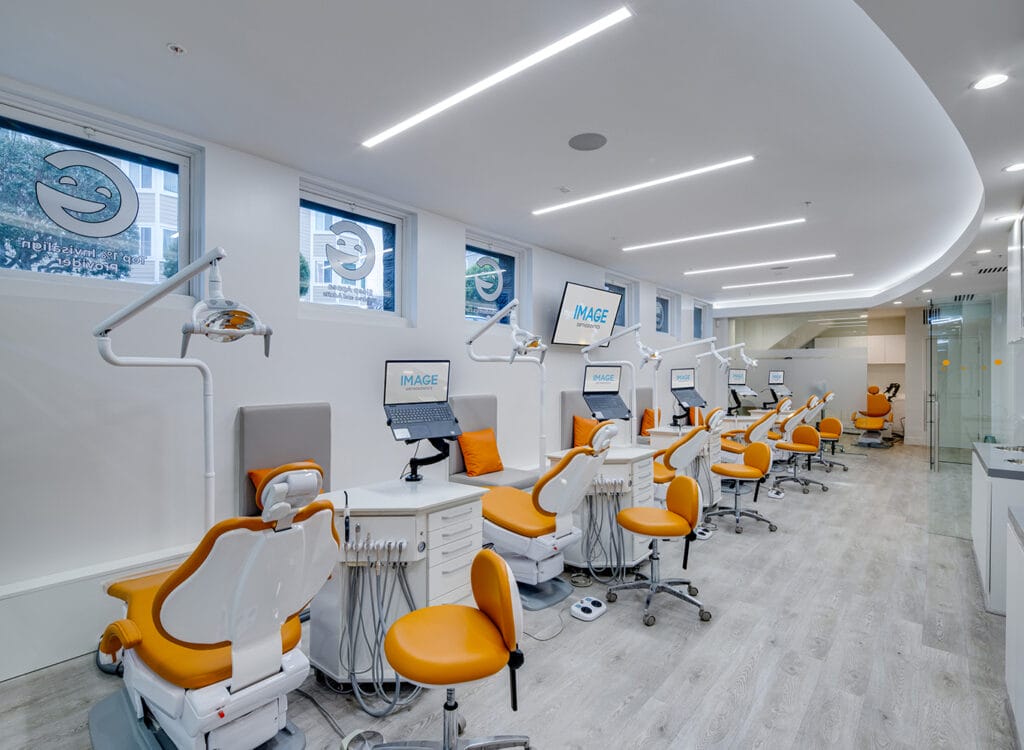 A modern dental clinic with multiple orange and white dental chairs, workstations, overhead lights, and wall-mounted monitors arranged in a clean, well-lit space.