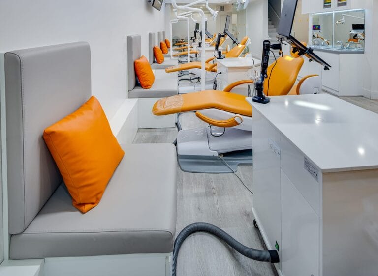 Row of gray bench seats with bright orange throw pillows alongside orange-and-white orthodontic chairs and overhead light arms in the Pacific Heights clinic treatment area.