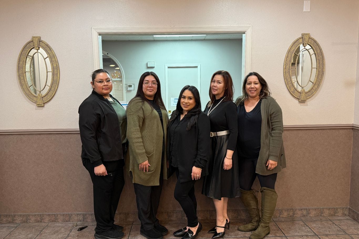 Five women stand indoors in front of a reception window, posing for a group photo. The background features two round mirrors on a beige wall.