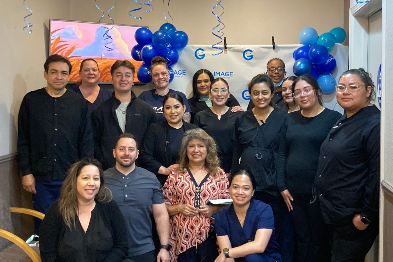 A group of sixteen adults pose together indoors in front of blue balloons and a banner, some wearing black or navy uniforms, at what appears to be a workplace event.