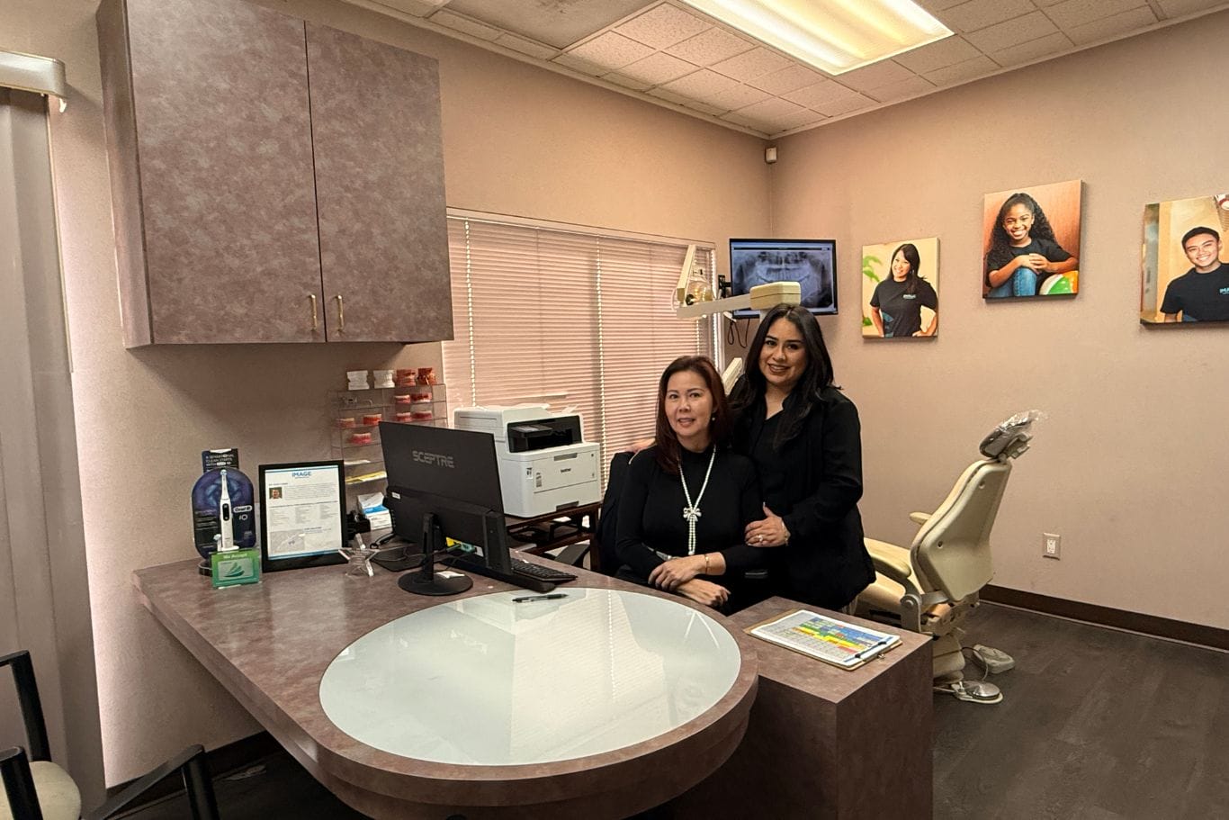 Two women in black attire pose in a dental office, one seated at a desk with a computer, the other standing beside her. Dental chair, equipment, and framed photos are visible in the background.