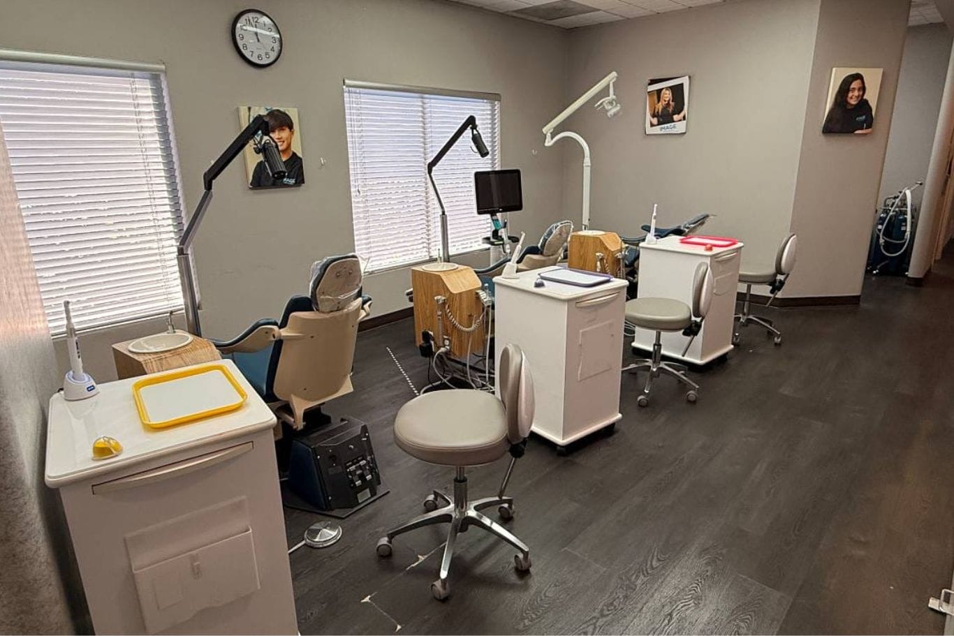Three dental exam stations with chairs, equipment, and desks are lined up in a clinic room with large windows, wall photos, and a clock.