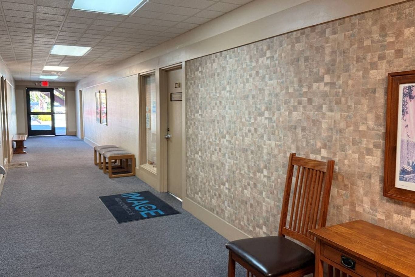 A carpeted hallway with beige walls, wooden benches, a chair, a small table, a framed picture, and a door mat labeled "MAGEE" near an office door.