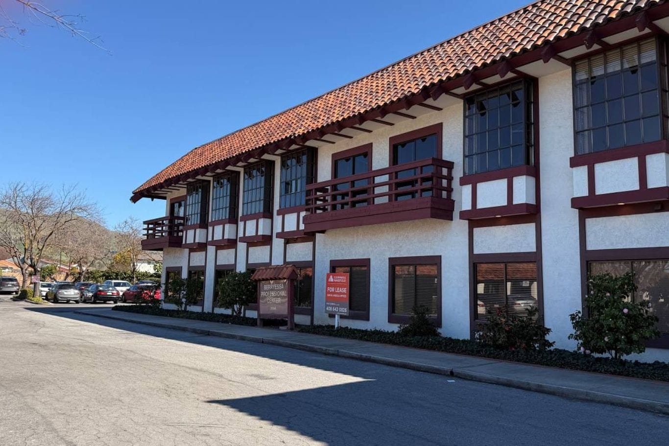 Two-story building with white walls and red trim, featuring large windows and a "For Lease" sign out front, set on a quiet street with several parked cars.