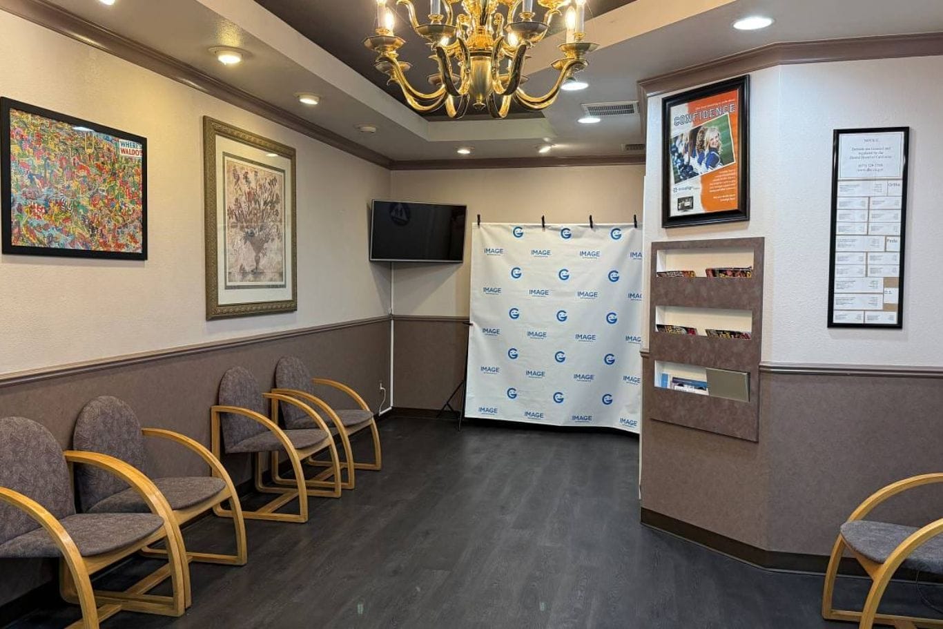 A waiting room with gray chairs, framed artwork on the walls, a TV, a chandelier, brochure racks, and a corner banner with the PMG logo.