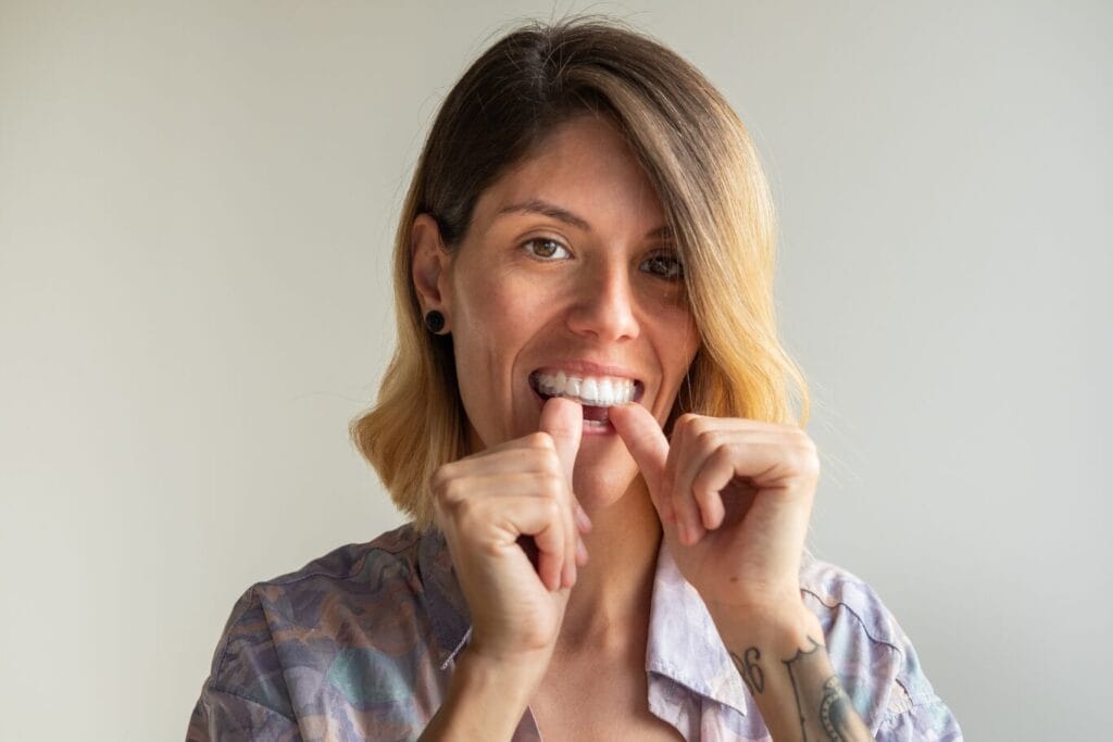 A person with shoulder-length hair is smiling while putting on a clear dental aligner, using both hands to fit it onto their upper teeth, wondering, "Can Invisalign fix overbites?.