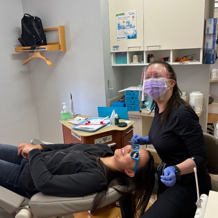 A dental hygienist at San Francisco Post Street Orthodontist, wearing PPE, stands beside a smiling patient in a dental chair.
