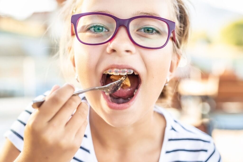 Close-up of a smiling child wearing braces, holding a fork and taking a bite of food