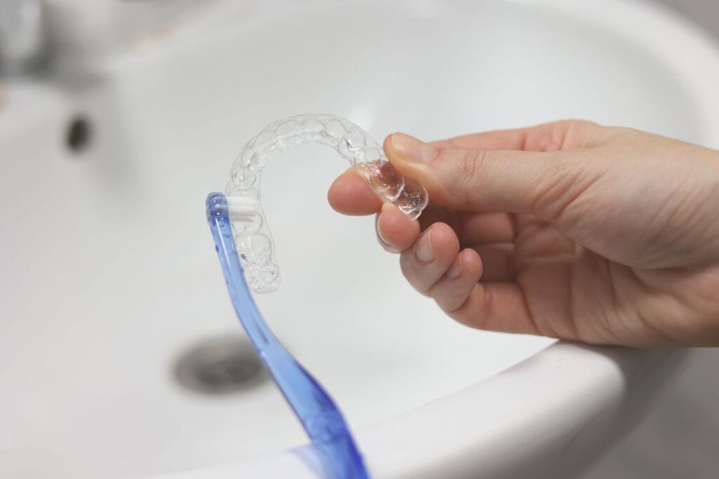 Hand holding a clear dental aligner while rinsing it at a bathroom sink with a toothbrush
