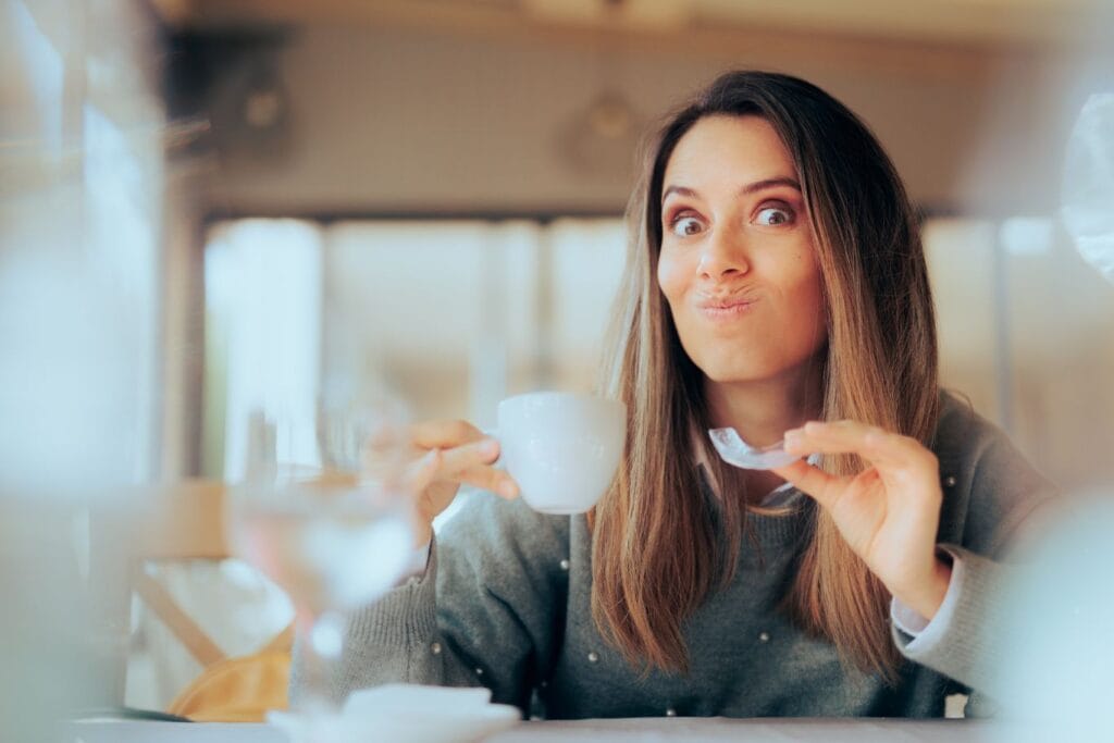 Follow us for more tips like, how to clean invisalign aligners. Woman making a playful expression while holding a small cup of coffee and a clear aligner