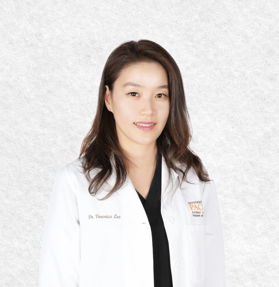 A woman with long dark hair wears a white lab coat embroidered with "Dr. Veronica Lee." As a Post Street Orthodontist, she specializes in Invisalign and clear aligners, standing confidently against a plain light background.