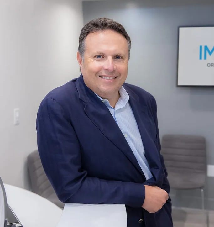A man in a blue blazer and light shirt stands indoors, smiling at the camera with one arm resting on a white counter, in what appears to be a San Francisco Pacific Heights orthodontist office. Gray chairs and a sign are visible in the background.