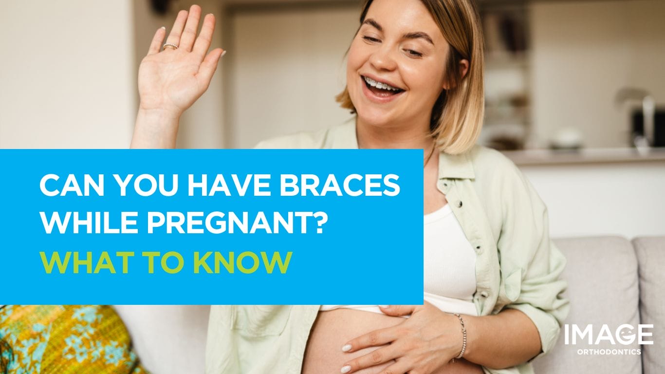 Smiling pregnant woman sitting on a sofa, wearing braces and waving