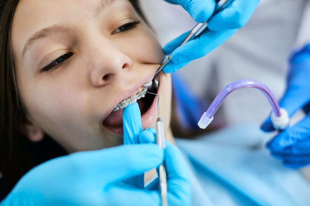 A dental professional adjusts braces on a patient’s teeth using dental tools while another uses a suction device, often prompting patients to wonder how long does the soreness of braces last after such adjustments.