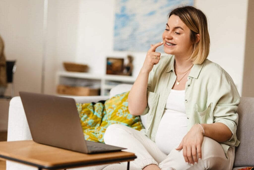 A pregnant woman sits on a couch, smiling and pointing to her braces while video chatting on a laptop in a living room.