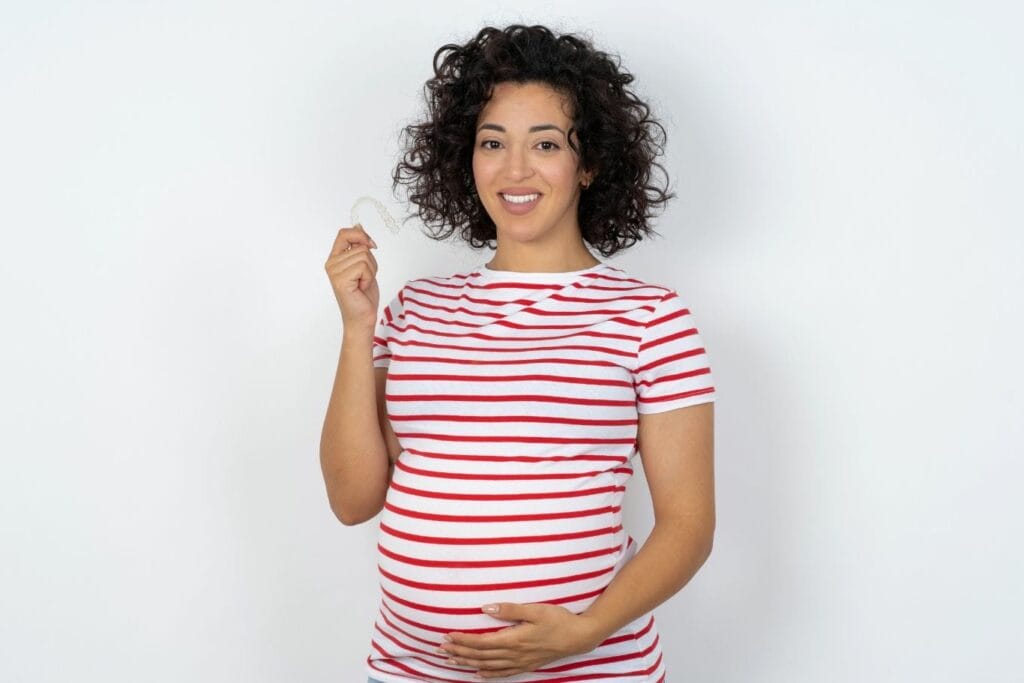 Smiling pregnant woman holding a clear Invisalign aligner in one hand and resting the other hand on her baby bump