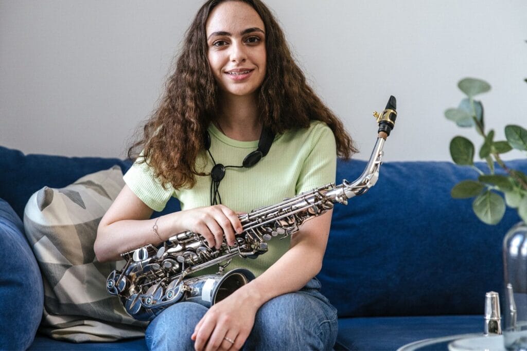 Young woman wearing braces sitting on a couch, holding a saxophone and smiling at the camera