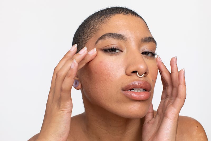 Person with short hair and a septum piercing touches their face with both hands, as if experiencing jaw pain, looking directly at the camera against a plain white background.