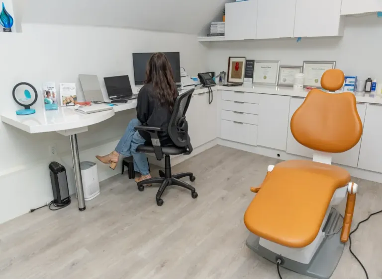 A person sitting at a desk with a computer in a dental office next to an orange exam chair.