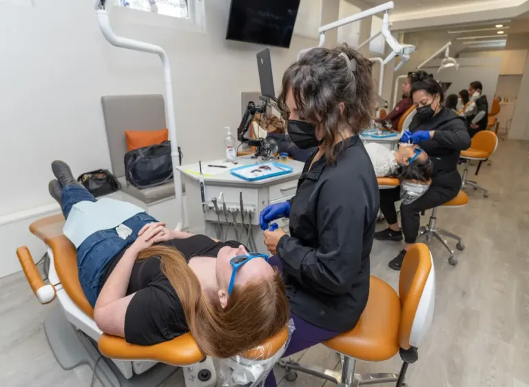 A dental hygienist in a mask and gloves treats a patient reclining in a chair, with other patients and staff visible in the background of the clinic.